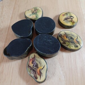 May include: A collection of small, vintage black boxes in various shapes, including round, oval, and heart-shaped. Some boxes feature painted lids with bird and floral designs. The boxes are arranged on a light wooden surface.