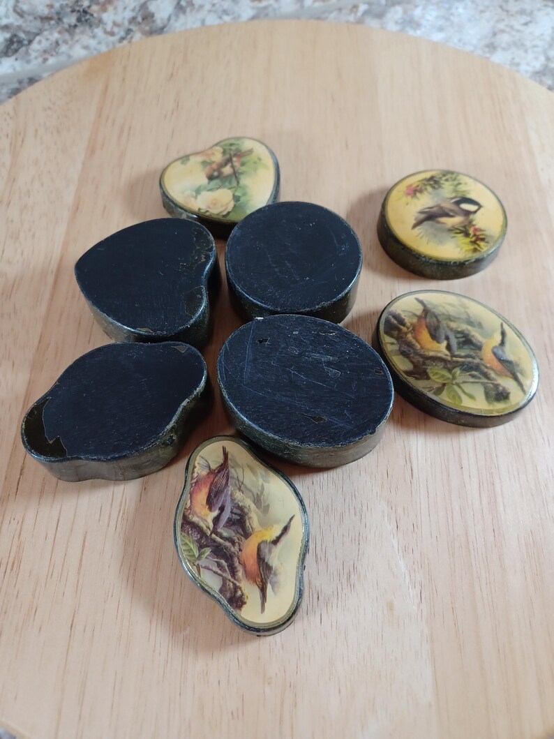 May include: A collection of small, vintage black boxes in various shapes, including round, oval, and heart-shaped. Some boxes feature painted lids with bird and floral designs. The boxes are arranged on a light wooden surface.