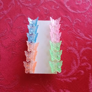 May include: A set of colorful butterfly hair clips in shades of blue, pink, orange, and green. The clips are arranged on a white card, showcasing their translucent design. These accessories are perfect for adding a touch of whimsy to any hairstyle.