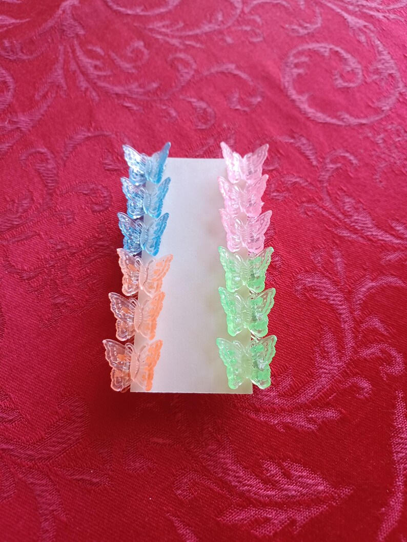 May include: A set of colorful butterfly hair clips in shades of blue, pink, orange, and green. The clips are arranged on a white card, showcasing their translucent design. These accessories are perfect for adding a touch of whimsy to any hairstyle.