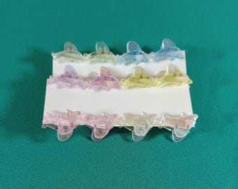 May include: A set of twelve small, translucent dolphin-shaped hair clips in various pastel colors, including pink, yellow, blue, and purple. The clips are arranged on a white card against a teal green background.