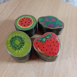 May include: Four small wooden boxes with different fruit designs. The boxes are painted with a watermelon, kiwi, strawberry, and a cluster of purple berries.