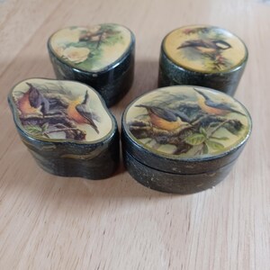May include: Four small, decorative boxes with lids. The boxes are dark green with a textured appearance. Each lid features a different bird illustration in a naturalistic style, with a yellow and green background. The boxes are arranged on a light brown surface.