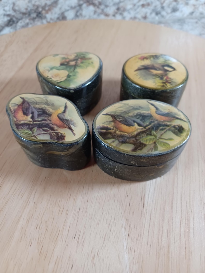 May include: Four small, decorative boxes with lids. The boxes are dark green with a textured appearance. Each lid features a different bird illustration in a naturalistic style, with a yellow and green background. The boxes are arranged on a light brown surface.