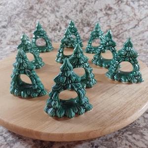 May include: A collection of nine ceramic Christmas tree-shaped napkin rings. The napkin rings are a glossy, deep green color and have a hole in the center. They are arranged on a round, light-colored wooden surface.