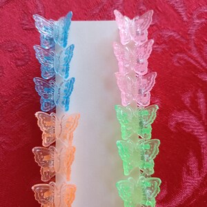 May include: A set of colorful butterfly hair clips in blue, pink, orange, and green. The clips are arranged in rows on a white card, with three clips of each color. The background is a red textured surface.