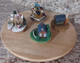 May include: A wooden serving tray displays four miniature decorative figurines. The figurines include a house with snow, a basket of fruit, a small house with a blue roof, and a barn with a silo. The figurines are on small wooden bases.