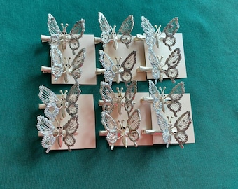 May include: Silver-toned butterfly hair clips arranged on small, rectangular cards. The butterfly wings have intricate detailing. The clips are displayed on a green surface. The butterflies are a decorative hair accessory.