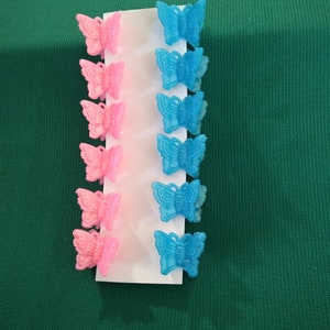 May include: A set of six butterfly hair clips in pink and blue. The butterfly clips are arranged on a white card, with three pink clips on the left and three blue clips on the right. The background is a solid green.