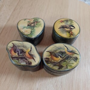 May include: Four small, vintage trinket boxes with bird illustrations. The boxes are black with decorative lids featuring various bird and floral designs. One box is heart-shaped, while the others are round or oval.