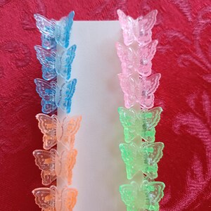 May include: A set of colorful butterfly hair clips in shades of blue, pink, orange, and green. The clips are arranged in rows on a white card, perfect for adding a touch of whimsy to any hairstyle. The background is a red fabric.