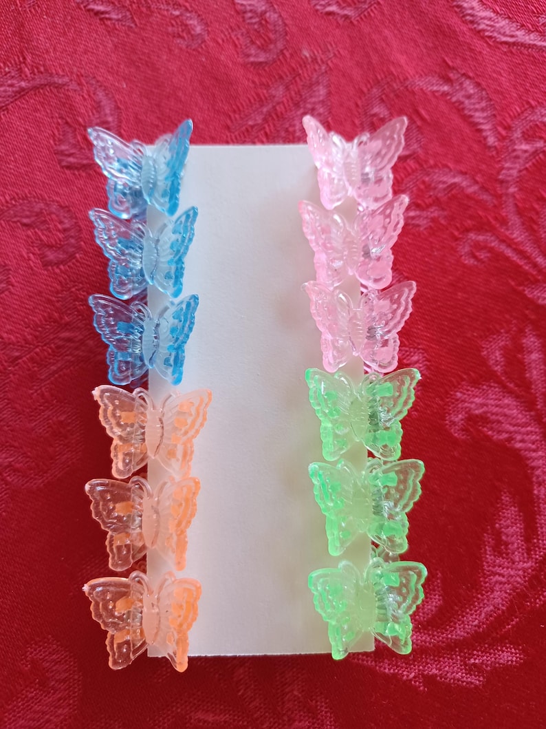 May include: A set of colorful butterfly hair clips in shades of blue, pink, orange, and green. The clips are arranged in rows on a white card, perfect for adding a touch of whimsy to any hairstyle. The background is a red fabric.