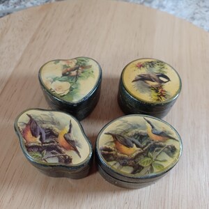 May include: Four small, vintage trinket boxes with bird illustrations. The boxes are black with lids featuring different bird and floral designs. One is heart-shaped, another round, and two are oval. The lids have a glossy finish.