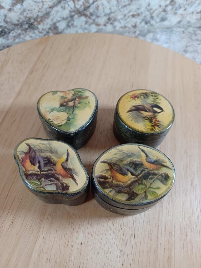 May include: Four small, vintage trinket boxes with bird illustrations. The boxes are black with lids featuring different bird and floral designs. One is heart-shaped, another round, and two are oval. The lids have a glossy finish.