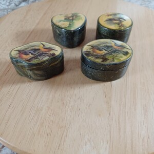 May include: Four small, antique trinket boxes with bird illustrations on the lids. The boxes are dark green with a gold trim and sit on a wooden surface. The bird illustrations feature various colors, including orange, yellow, and green.