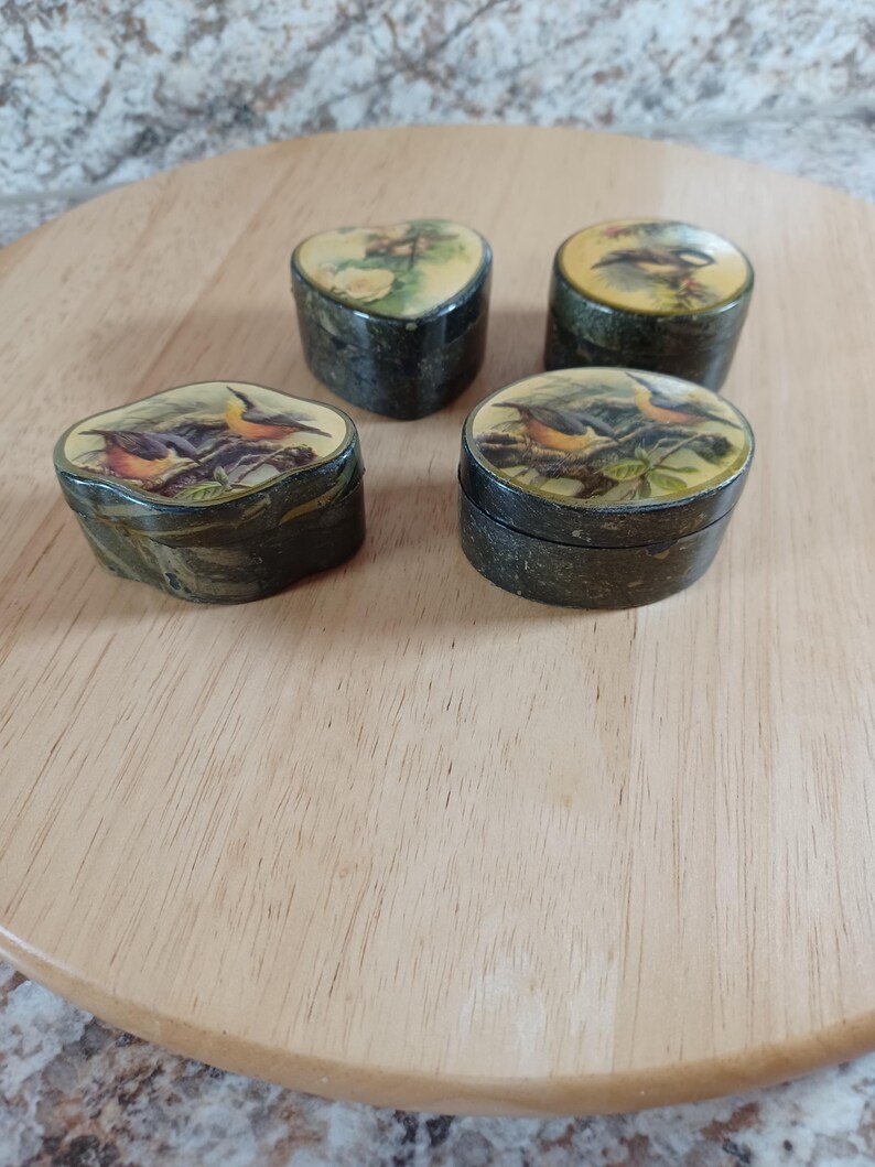 May include: Four small, antique trinket boxes with bird illustrations on the lids. The boxes are dark green with a gold trim and sit on a wooden surface. The bird illustrations feature various colors, including orange, yellow, and green.