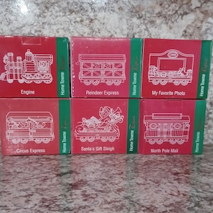 May include: Six red and white boxes with images of miniature train cars. The boxes are labeled with the names of the train cars: Engine, Reindeer Express, My Favorite Photo, Circus Express, Santa's Gift Sleigh, and North Pole Mail. Each box has the words "Home Towne" printed on the side.