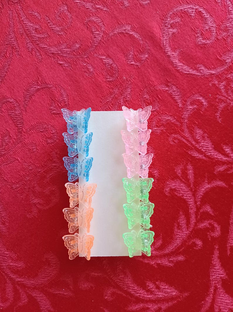 May include: A collection of small butterfly hair clips in various colors, including blue, pink, orange, and green. The clips are arranged on a white card against a red background. These accessories are ideal for adding a touch of whimsy to any hairstyle.