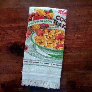 May include: A vintage white and green kitchen towel with a colourful illustration of a bowl of corn flakes and various fruits. The towel has the words "Kellogg's CORN FLAKES" and "FRUITS IN SEASON" printed on it, with a fringed bottom.