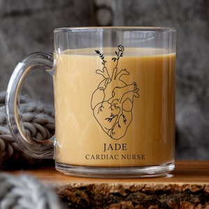 May include: A clear glass mug with a handle filled with a light brown beverage. The mug has a black and white illustration of a heart with a floral design and the text "JADE CARDIAC NURSE".