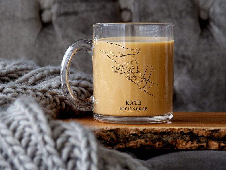 May include: A clear glass mug with a black outline of two hands holding each other. The mug has the text "KATE NICU NURSE" printed on it. The mug is filled with a light brown liquid.