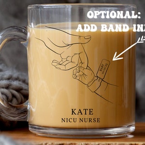 May include: A clear glass mug with a handle, filled with a light brown liquid. The mug has a black line drawing of two hands holding each other, with the text "KATE NICU NURSE" below. A small tag with the text "BABY 01/01/23" is on the wrist of the smaller hand.