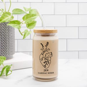 May include: A clear glass tumbler with a bamboo lid and a glass straw. The tumbler features a black line drawing of a heart with floral accents and the text "JEN CARDIAC NURSE". The glass is filled with a light brown beverage.
