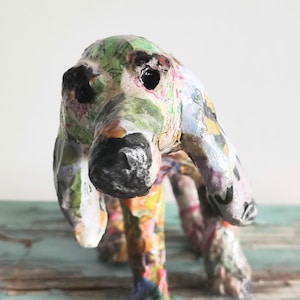 May include: A colorful, handcrafted dog sculpture. The dog has a black nose and eyes, with a body covered in a collage of patterned paper in shades of green, pink, orange, and blue. The sculpture is standing on a light blue surface.