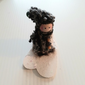 Puede incluir: Una pequeña muñeca hecha a mano que lleva un gorro y un abrigo de punto negro y gris, de pie sobre una base blanca en forma de corazón con purpurina.