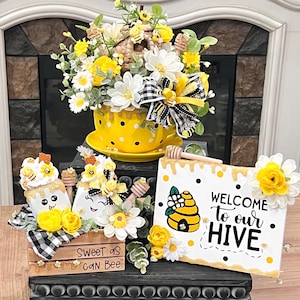 May include: A yellow and black bee-themed home decor set with a floral arrangement in a yellow teacup, two white and yellow bee-shaped figurines, and a wooden sign that reads "Welcome to our Hive".
