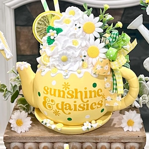 May include: A yellow teapot with the words "sunshine & daisies" and white polka dots. The teapot is decorated with white daisies, lemon slices, and green accents. A decorative piece for the kitchen or dining room.