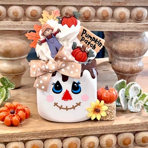 May include: A white ceramic container with a smiling face design, decorated with a bow, pumpkins, and a scarecrow. The container has a "Pumpkin Patch" sign and is surrounded by fall-themed decorations, including a sunflower and fall leaves.