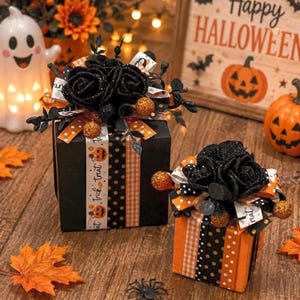 May include: Two square gift boxes decorated with black glittery roses, orange and black ribbons, and small glittery spheres. The larger box is black, and the smaller box is orange. Both feature ribbons with a "Trick or Treat" text print.