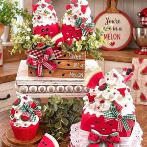 May include: Decorative watermelon-themed figurines with whipped cream, watermelon slices, and bows. The figurines are red with smiling faces and are arranged with greenery and a wooden crate that says "Sweet as a Melon."