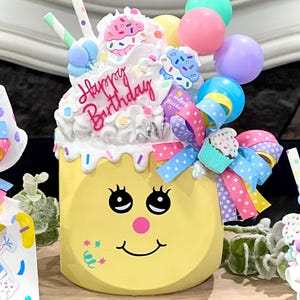 May include: A yellow, cake-shaped decoration with a smiling face, topped with white frosting, sprinkles, and colorful balloons. The words "Happy Birthday" are written in pink script. A decorative bow and straws complete the festive design.