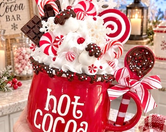 May include: A red ceramic mug with the text hot cocoa in white letters, topped with faux whipped cream, chocolate squares, red and white peppermint candies, and white snowflakes. A matching spoon with a red and white striped handle is attached.