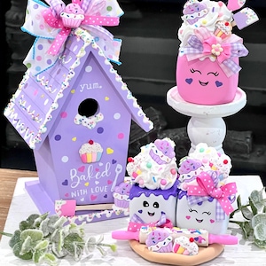 May include: Purple birdhouse with a pink bow and the text "Baked with Love" and a cupcake design.  Two pink and purple marshmallow-like figures with faces and sprinkles.  A pink cupcake with a white frosting and sprinkles on a white pedestal.