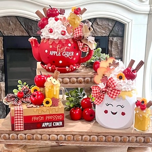 May include: A decorative fall-themed display featuring a red teapot and a white mug, both adorned with whipped cream, apples, and cinnamon sticks. The teapot reads "Hot Apple Cider". A box reads "Fresh Pressed Apple Cider".