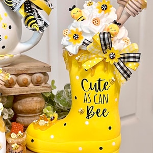 May include: Yellow rain boot with white polka dots and black dots. The boot has a white and black checkered ribbon with a yellow bow. The boot is decorated with bees, flowers, and honeycombs. The boot has the text "Cute as can Bee" written on it.