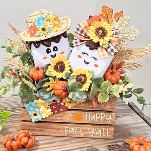 May include: A wooden crate decorated for fall, featuring two marshmallow-shaped figures with smiling faces, adorned with hats and surrounded by sunflowers, pumpkins, and foliage. The crate has the text "Happy Fall y'all".