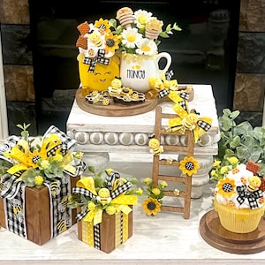 May include: A collection of bee-themed decorative items, including a yellow cup with "HONEY" text, a wooden ladder, and gift boxes. The items feature sunflowers, bees, and black and white checkered ribbons. A cupcake and a tray with bee decorations are also present.