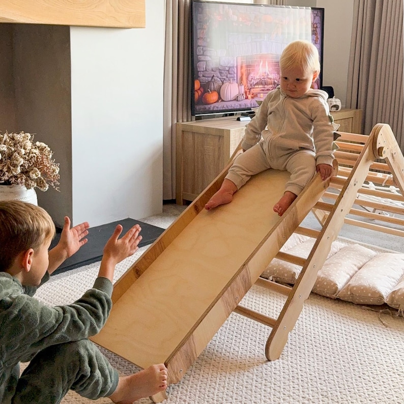 May include: A wooden climbing triangle with a slide. The slide is made of light-colored wood and is angled down. A child in a beige jumpsuit sits at the top of the slide. Another child in a green jumpsuit is at the bottom.