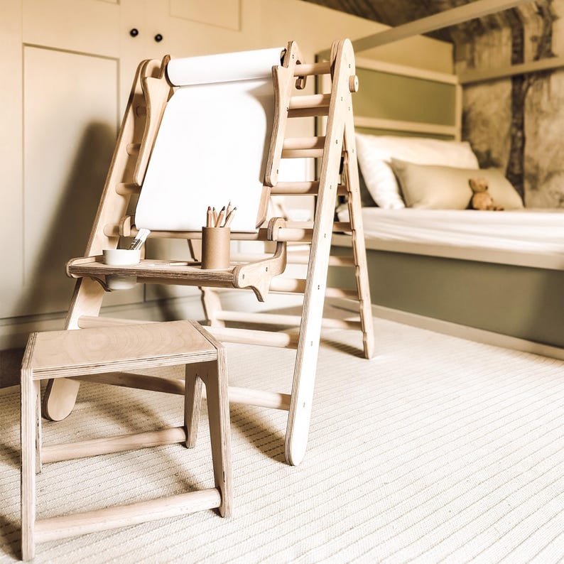May include: A wooden children's art easel with a roll of white paper, a small stool, and a shelf for art supplies. The easel has a ladder-like design and is made of light-colored wood. Pencils and a small bowl are on the shelf.