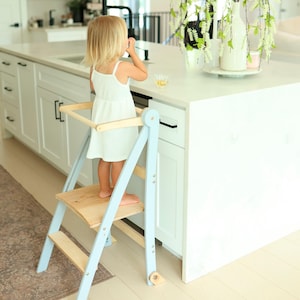 May include: A light blue and natural wood learning tower with a safety rail and two steps. The structure features a sturdy frame with a wide platform for standing, designed to provide elevation for reaching kitchen counters safely and securely.