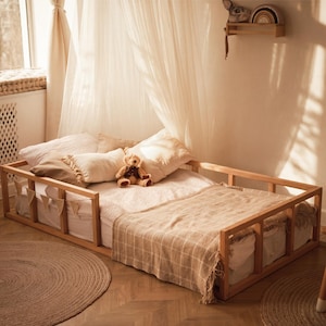 May include: A low-profile wooden floor bed frame with a rectangular structure and safety rails. The bed is dressed with white bedding, a beige plaid throw blanket, and several pillows, including a small plush teddy bear sitting in the center.