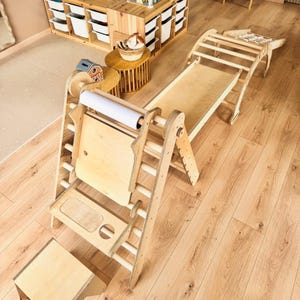 May include: Wooden climbing structure with a slide, ladder, and drawing board. The natural wood-toned play set includes a slide, a climbing arch, and a drawing board with a roll of white paper. A small wooden stool is also included.