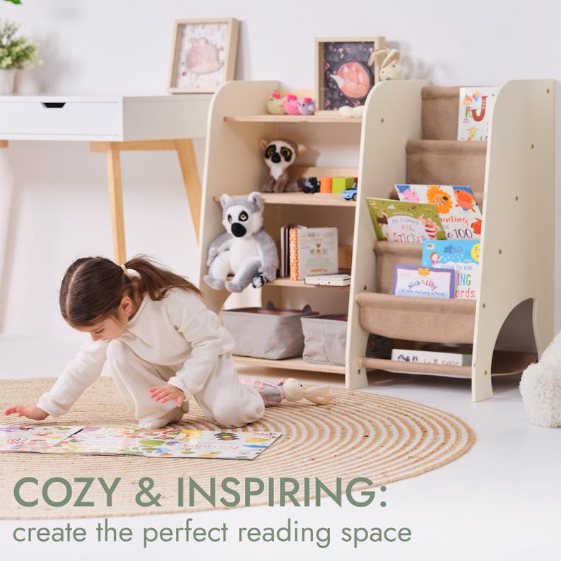 May include: A child's reading nook featuring two cream-colored bookshelves filled with books and stuffed animals. The shelves have a unique stepped design with fabric pockets. A young girl is playing on a round jute rug in front of the shelves. Text reads: COZY & INSPIRING: create the perfect reading space.