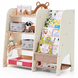 May include: A cream-colored wooden bookshelf with multiple shelves, holding children's books, toys, and storage bins. The bookshelf has a unique design with angled sides and a fabric book holder. Toys include a stuffed bunny and a lemur.