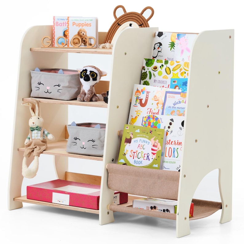 May include: A cream-colored wooden bookshelf with multiple shelves, holding children's books, toys, and storage bins. The bookshelf has a unique design with angled sides and a fabric book holder. Toys include a stuffed bunny and a lemur.