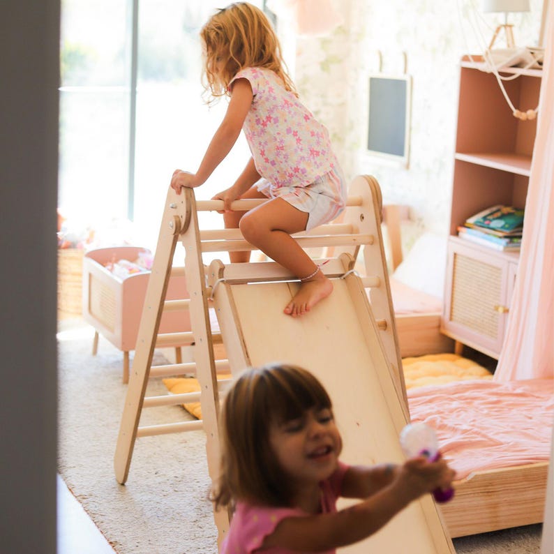 May include: A wooden climbing arch with a slide, designed for children's play. The arch is light-colored wood, and the slide is a smooth, flat surface. A child is climbing on the arch, while another child is playing in the foreground. The room has a pink theme.